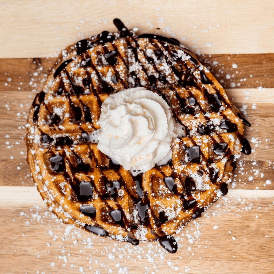 A golden waffle drizzled with chocolate sauce, powdered sugar, and topped with whipped cream.