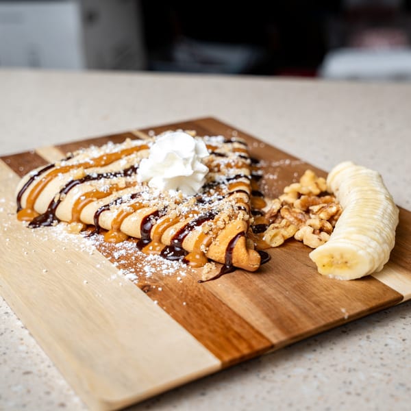 Crepe D'France with chocolate, caramel, whipped cream, walnuts, and sliced banana on a wooden board.
