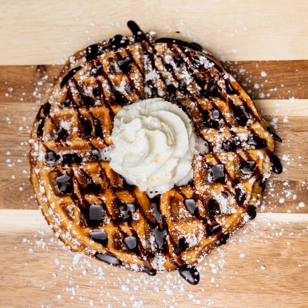 A golden waffle with chocolate drizzle, powdered sugar, and whipped cream on a wooden surface.