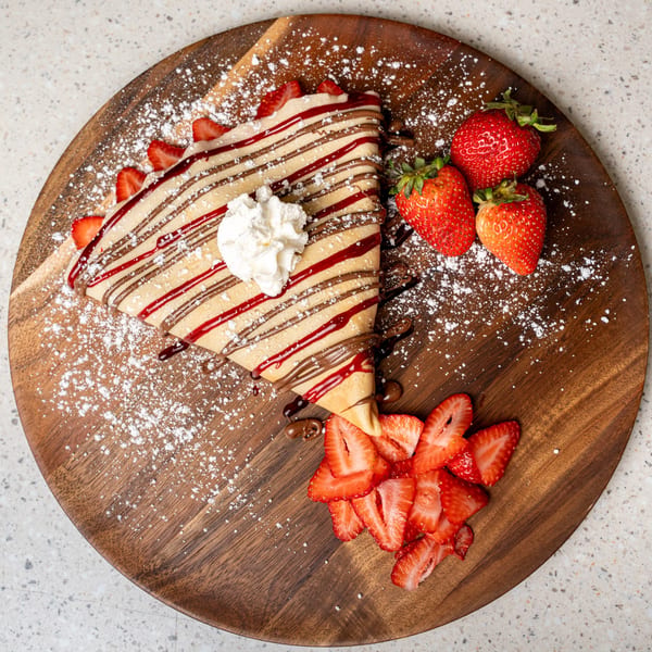 A strawberry and chocolate crepe with whipped cream, powdered sugar, and fresh strawberries on a wooden board.