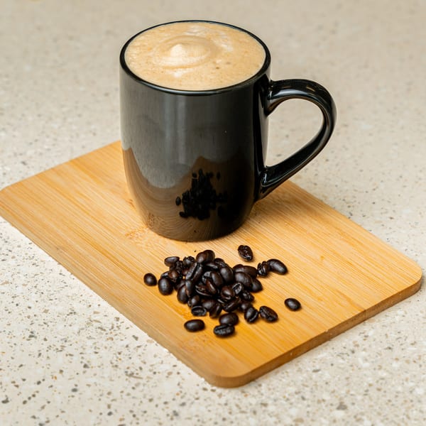 Black mug of frothy coffee and scattered coffee beans on a wooden board.
