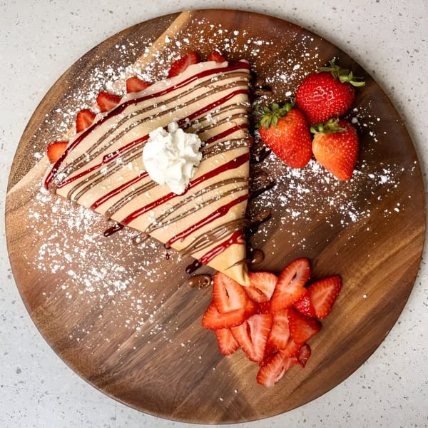 A strawberry and chocolate drizzled crepe with whipped cream and fresh strawberries on a wooden board.